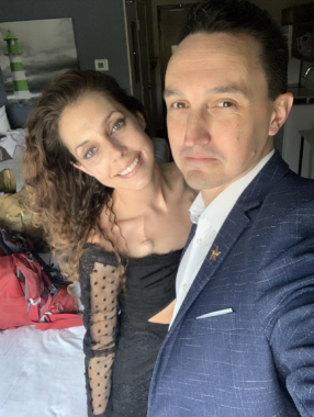 A man and woman pose for a selfie while dressed in formal attire. The man is wearing a blue suit jacket over a white collared shirt and holding his left arm out, presumably to take the photo. The woman is standing just behind him wearing a black dress with mesh sleeves. They appear to be standing inside their home, perhaps in a bedroom.